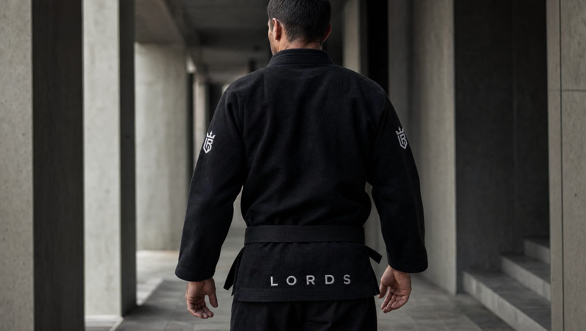 Atleta de costas caminhando por corredor de concreto, kimono preto LORDS com faixa preta